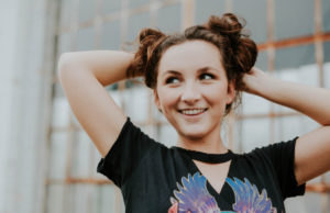 Brunette young woman with her hair in two top buns and her hands behind her head looks off to the side and smiles with dental crowns