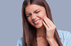 Brunette young woman cringes in pain and touches her hand to her cheek due to impacted wisdom teeth