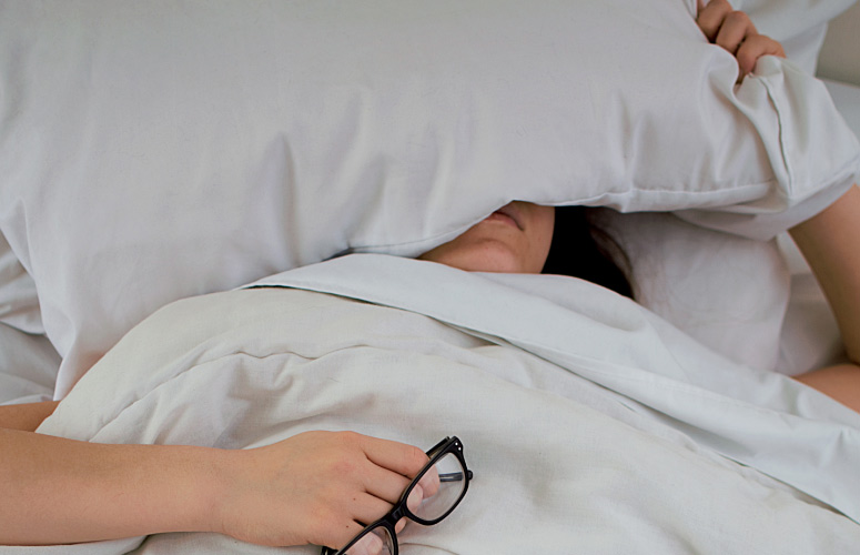 woman holding eye glasses in her hand sleeping with a pillow over her head